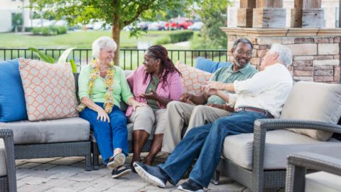 A group of senior friends hanging out together, sitting outdoors on a patio conversing. The African-American couple are in their 50s. The Caucasian couple are in their 70s. The women are sitting next to each other, talking, and the men are chatting with each other.