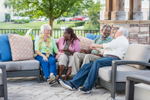A group of senior friends hanging out together, sitting outdoors on a patio conversing. The African-American couple are in their 50s. The Caucasian couple are in their 70s. The women are sitting next to each other, talking, and the men are chatting with each other.