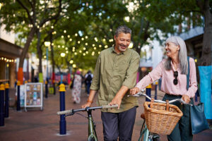 Smiling old couple walking with bicycles in the street as they enjoy the benefits of positivity and healthy living. Happy senior multiethnic man and beautiful mature woman enjoying a bike ride together in town. Multiethnic elderly couple on a romantic evening walk laughing while pushing their bike in a pedestrian area.