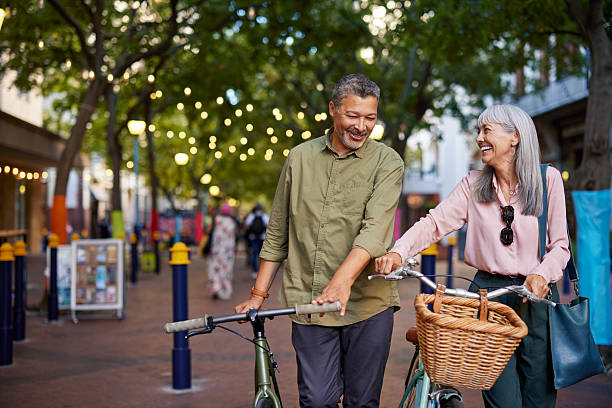 Smiling old couple walking with bicycles in the street as they enjoy the benefits of positivity and healthy living. Happy senior multiethnic man and beautiful mature woman enjoying a bike ride together in town. Multiethnic elderly couple on a romantic evening walk laughing while pushing their bike in a pedestrian area.